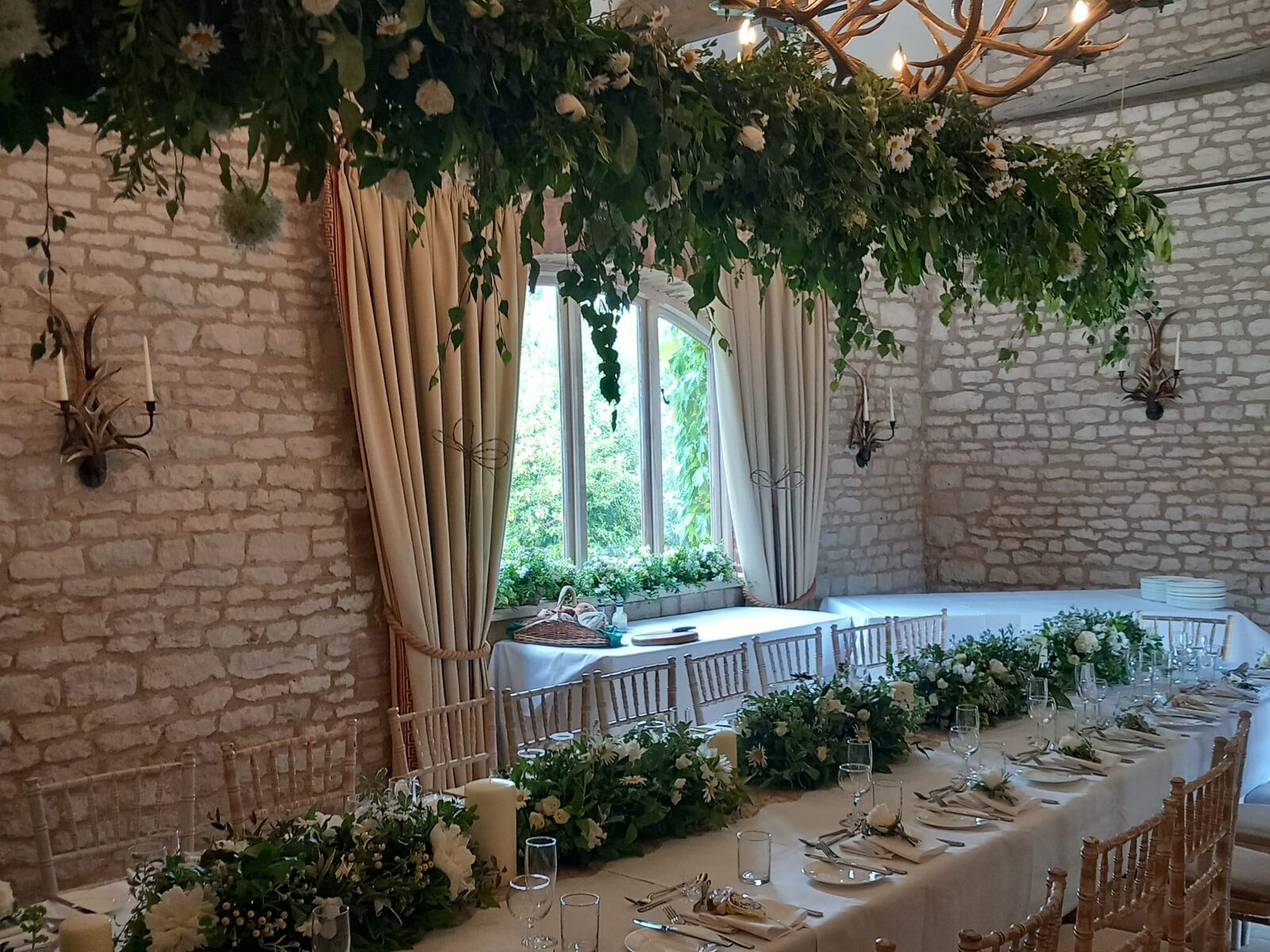 A long table laid by Penny Mardon Caters....: white linen and silver beneath a running garland of white flowers and foliage, in a stone barn