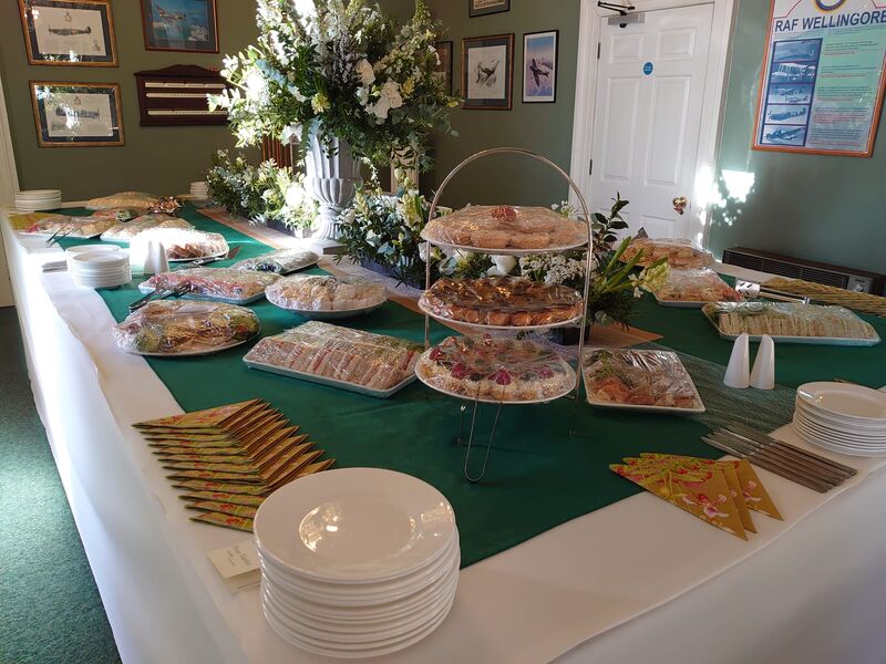 A village hall buffet with a green runner, tiered pastries and a tall white floral urn