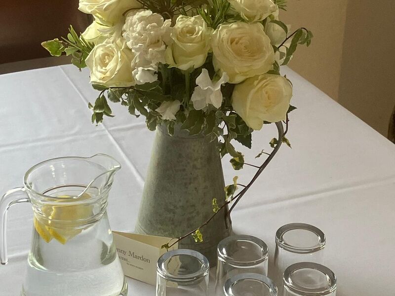 White roses and foliage in a galvanised jug beside a water jug and tumblers